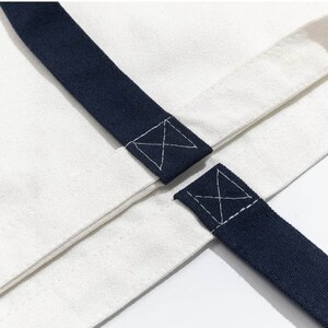 May include: Close-up of a white canvas tote bag with navy blue straps. The straps are sewn to the bag with an X-shaped stitch.