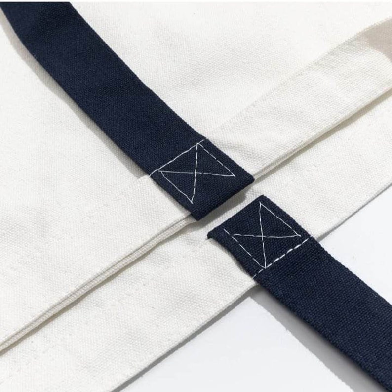 May include: Close-up of a white canvas tote bag with navy blue straps. The straps are sewn to the bag with an X-shaped stitch.