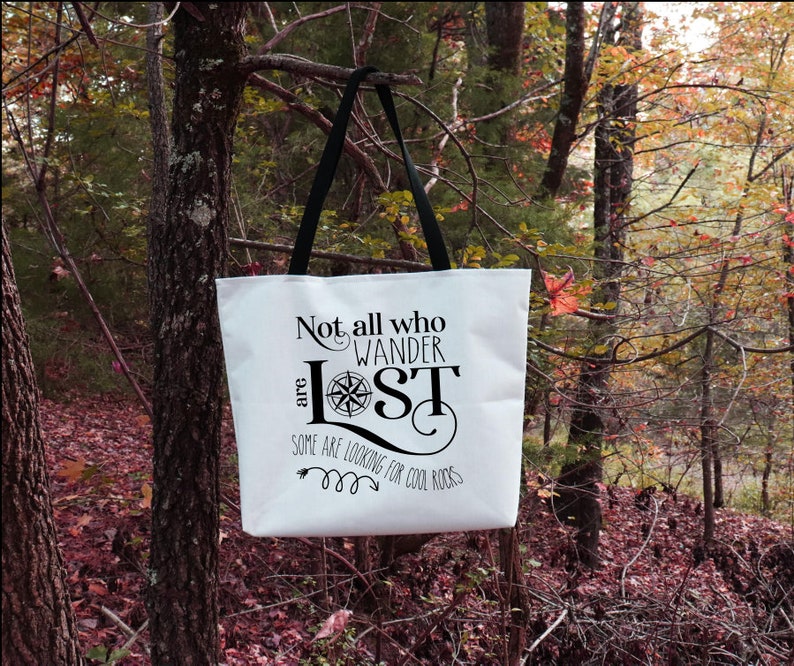 May include: White canvas tote bag with black straps and a black graphic that reads "Not all who wander are lost. Some are looking for cool rocks."