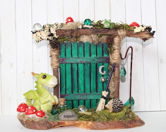 May include: A miniature fairy house with a green door, a small dragon figurine, a hedgehog figurine, and a sign that says "Dream". The house is made of twigs and moss and has a whimsical, magical feel.