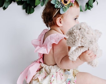 May include: A baby girl wearing a pink and white ruffled dress with a floral headband and holding a plush toy.