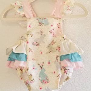 May include: A white baby onesie with a floral and bunny print. The onesie has a pink, white, and blue ruffle trim around the legs and arms.