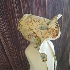 May include: A white bonnet with a colorful print featuring characters from the book "Where the Wild Things Are". The bonnet has a ruffle trim and ties at the back.