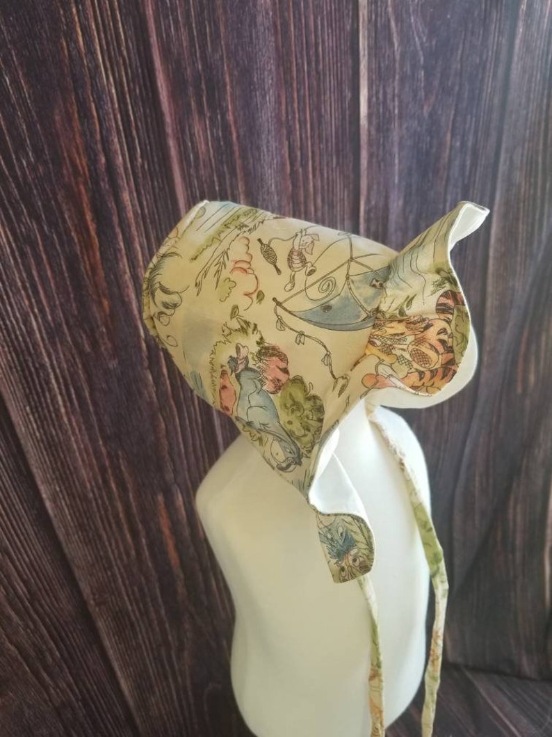May include: A white bonnet with a colorful print featuring characters from the book "Where the Wild Things Are". The bonnet has a ruffle trim and ties at the back.