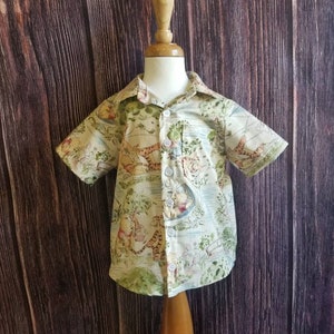 May include: A short-sleeved button-down shirt for a child, featuring a light beige background with a colorful print of characters from the book "The Wind in the Willows".