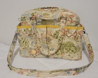 May include: A beige canvas tote bag with a yellow trim and a Winnie the Pooh print. The bag has a long adjustable strap and two top handles.