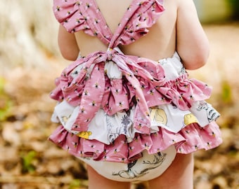 May include: A pink and white baby dress with a bee print and a ruffled skirt. The dress has a criss-cross back with a bow.