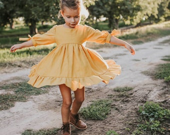 May include: A young girl wearing a yellow dress with a ruffled hem and brown boots spins around on a dirt path.
