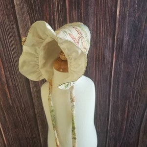 May include: A white bonnet with a floral print featuring a variety of colors, including green, pink, and yellow. The bonnet has a ruffle around the edge and ties that can be tied under the chin.