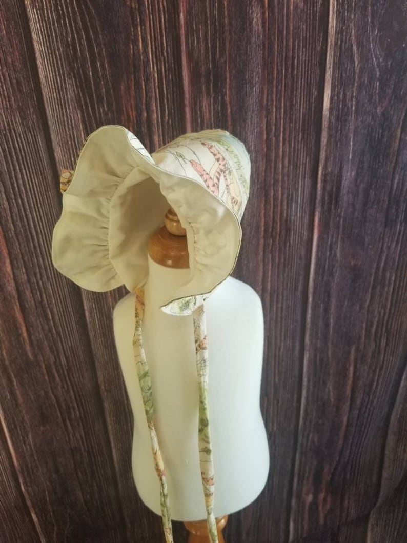 May include: A white bonnet with a floral print featuring a variety of colors, including green, pink, and yellow. The bonnet has a ruffle around the edge and ties that can be tied under the chin.
