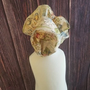 May include: A cream-colored baby bonnet with a ruffled edge and a whimsical print of a map with birds, flowers, and trees.