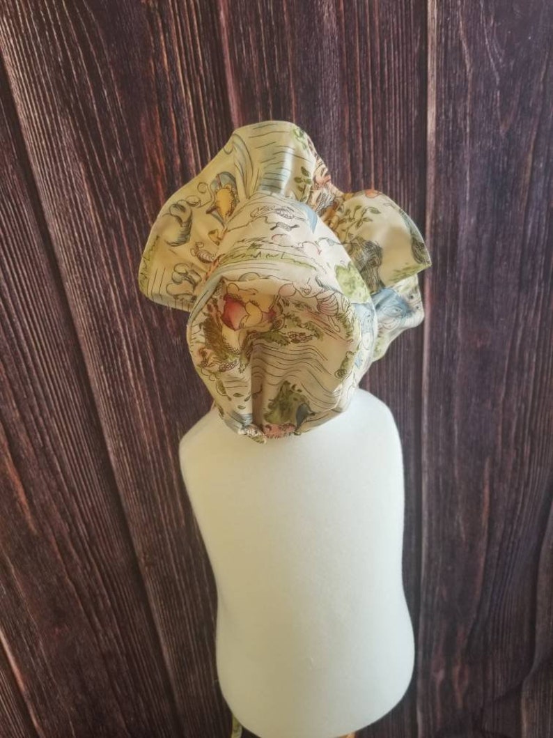 May include: A cream-colored baby bonnet with a ruffled edge and a whimsical print of a map with birds, flowers, and trees.