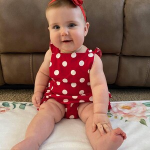 May include: A baby girl wearing a red and white polka dot dress with a red bow headband. She is sitting on a white blanket with a floral pattern.