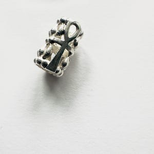 Peut inclure: Une perle argentée ornée d'un symbole Ankh. La perle cylindrique présente une surface texturée, avec l'Ankh au centre. Cette perle est probablement destinée à la fabrication de bijoux, et mesure environ 1 cm de long.