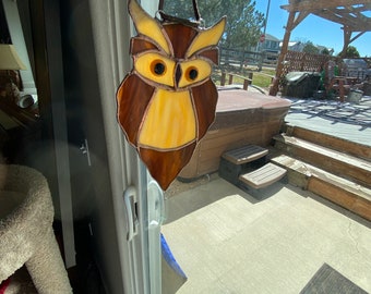 May include: A stained glass owl ornament with brown, yellow, and orange glass pieces. The owl has large, round, yellow eyes. The ornament is hanging from a brown leather cord.