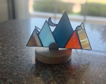 May include: A stained glass mountain range ornament featuring triangular panels in shades of orange, light blue, dark blue, and textured gold, with a small crescent moon shape, all mounted on a circular light-colored wooden base.