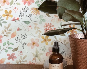 May include: A white ceramic tray with a gold bracelet, a brown glass bottle with a spray nozzle, and a silver smartwatch. The tray is on a wooden surface in front of a potted plant with green leaves and a white background with a floral pattern. The bottle has a label that reads "Botany Garden", "Concentrated Plant Room Spray".