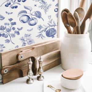 May include: A white background with a blue and white floral pattern featuring apples, cherries, berries, and leaves. The pattern is on a wall behind a wooden cutting board, two silver salt and pepper shakers, a white ceramic jar with a wooden lid, and a white ceramic dish with a gold spoon.
