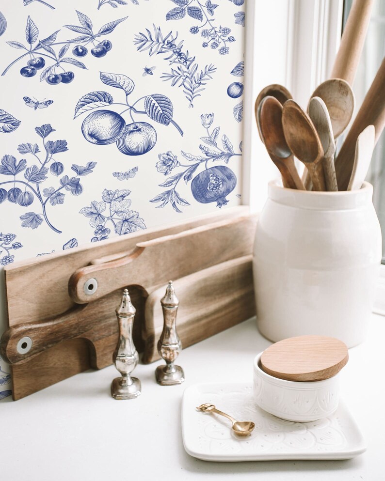 May include: A white background with a blue and white floral pattern featuring apples, cherries, berries, and leaves. The pattern is on a wall behind a wooden cutting board, two silver salt and pepper shakers, a white ceramic jar with a wooden lid, and a white ceramic dish with a gold spoon.