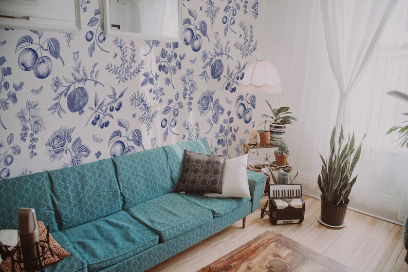 May include: A teal blue sofa with two pillows sits in a living room with white walls and a blue and white floral wallpaper. A potted plant is in the corner of the room.