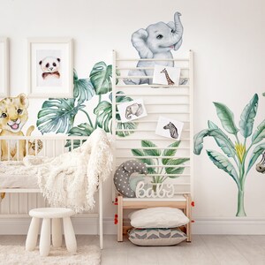 May include: A nursery room decorated with jungle animal wall decals. The decals feature a giraffe, lion, elephant, zebra, and palm trees. The decals are in a watercolour style and are a light blue, green, and brown colour.