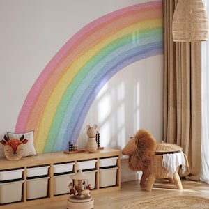 Könnte beinhalten: Kinderzimmer mit großem Wandtattoo in Regenbogenfarben. Darunter eine Holzaufbewahrung mit weißen Behältern. Dekoration: Kissen mit Hirschkopf, Wäschekorb in Löwenform aus Korbgeflecht und Holzkarussell.