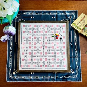Puede incluir: Un juego de mesa enmarcado con símbolos y texto rojos y negros, con piezas de juego coloridas. El juego está sobre una tela azul oscuro con un borde blanco. Una baraja de cartas del tarot y unas monedas están al lado del juego.