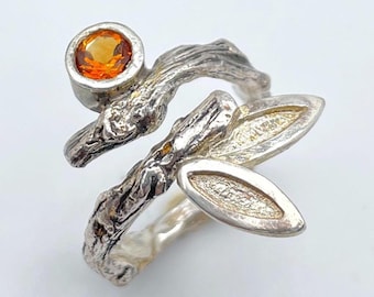 May include: A silver ring with a nature-inspired design. The ring features a textured band that resembles a tree branch, with a round orange gemstone and two leaf-shaped accents. The ring is set against a white background.
