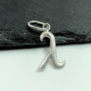 K&ouml;nnte beinhalten: Ein silberner Anh&auml;nger in Form des Buchstabens "R" mit geb&uuml;rsteter Oberfl&auml;che. Der Anh&auml;nger hat oben eine &Ouml;se zum Anbringen an einem Armband oder einer Halskette.