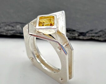 May include: A silver ring featuring a geometric, angular band design with a square-cut yellow gemstone set at the top. The metal surface has a matte, textured finish, and the structure includes visible rivets along the sides of the band.