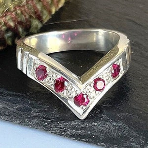 Könnte beinhalten: Ein silberner V-förmiger Ring mit fünf runden, roten Edelsteinen. Der Ring hat eine polierte Oberfläche und ist vor einem dunklen Hintergrund platziert. Das Design des Rings ist modern und geometrisch, geeignet für den Alltag oder besondere Anlässe.