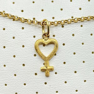 May include: A gold necklace with a heart-shaped pendant featuring a female symbol inside. The pendant has a textured finish.