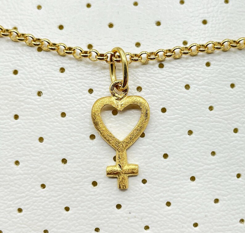 May include: A gold necklace with a heart-shaped pendant featuring a female symbol inside. The pendant has a textured finish.