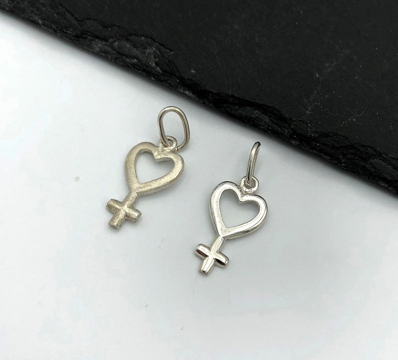 May include: Two silver charms in the shape of a heart with a female symbol inside. The charms are on a white background.