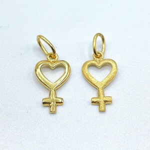 May include: Two gold-colored charms in the shape of a heart with a female symbol inside. The charms are on a white background.