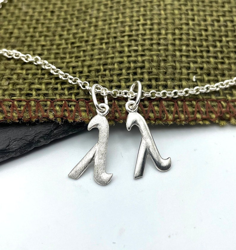 K&ouml;nnte beinhalten: Eine silberne Halskette mit zwei Anh&auml;nger in Form des Buchstabens 'A'. Die Anh&auml;nger sind strukturiert und haben eine geb&uuml;rstete Oberfl&auml;che.