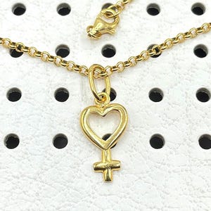 May include: A gold chain necklace with a heart-shaped pendant featuring a female symbol. The pendant is hollow and has a delicate design.