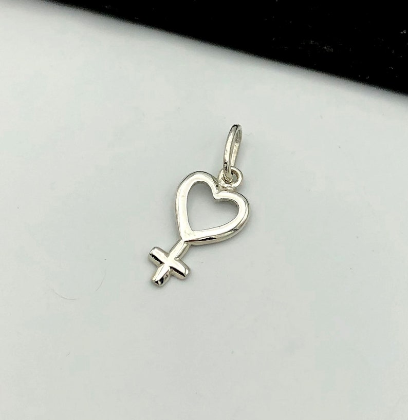 May include: A silver charm in the shape of a heart with a female symbol inside. The charm is designed to be worn on a necklace or bracelet.