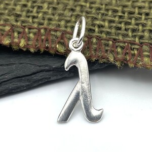 K&ouml;nnte beinhalten: Ein silberner Anh&auml;nger in Form des Buchstabens Lambda. Der Anh&auml;nger hat eine glatte, geb&uuml;rstete Oberfl&auml;che und eine &Ouml;se oben zum Anbringen an einer Halskette oder einem Armband.