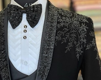 May include: A black tuxedo with intricate beaded embellishments. The jacket features a satin lapel and detailed floral patterns on the shoulders and sleeves. A black bow tie and a white dress shirt with a textured bib complete the formal ensemble.