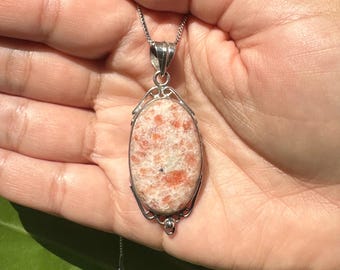 May include: A silver pendant necklace featuring an oval-shaped, peach-colored sunstone. The stone has orange and white flecks and is set in a decorative silver frame. The necklace is displayed against a green leaf background.
