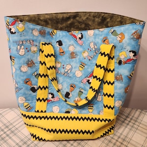 May include: A large tote bag featuring a light blue background with a repeating pattern of cartoon characters. The bag has yellow straps and a yellow base with a black zig-zag design. The interior lining is a dark green.