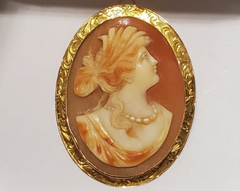 May include: A gold-toned oval cameo pendant featuring a carved portrait of a woman with a detailed hairstyle. The cameo is set in a gold frame with intricate floral designs.