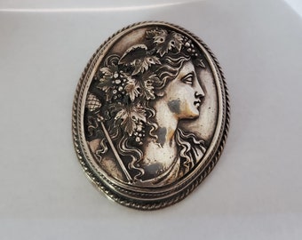 May include: A silver oval brooch with a detailed relief of a woman's profile with a crown of leaves and grapes.