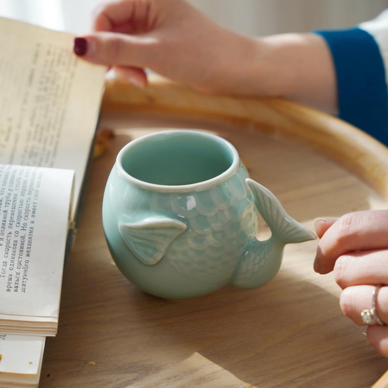 May include: A light blue ceramic mug shaped like a fish, with a tail and fin details. The mug is on a wooden surface. The mug has a round body and a handle shaped like a fish tail.