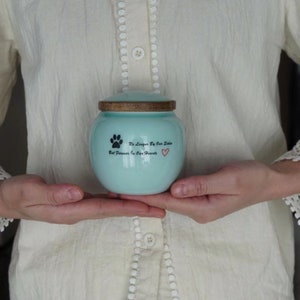 May include: A light blue ceramic pet urn with a wooden lid. The urn has a paw print and the text "No Longer By Our Side But Forever In Our Hearts" with a red heart.