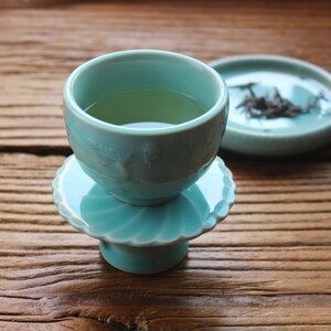 May include: A teal ceramic teacup filled with green tea, resting on a matching saucer with a scalloped edge. A second saucer with tea leaves sits nearby. The teacup and saucers are on a wooden surface.