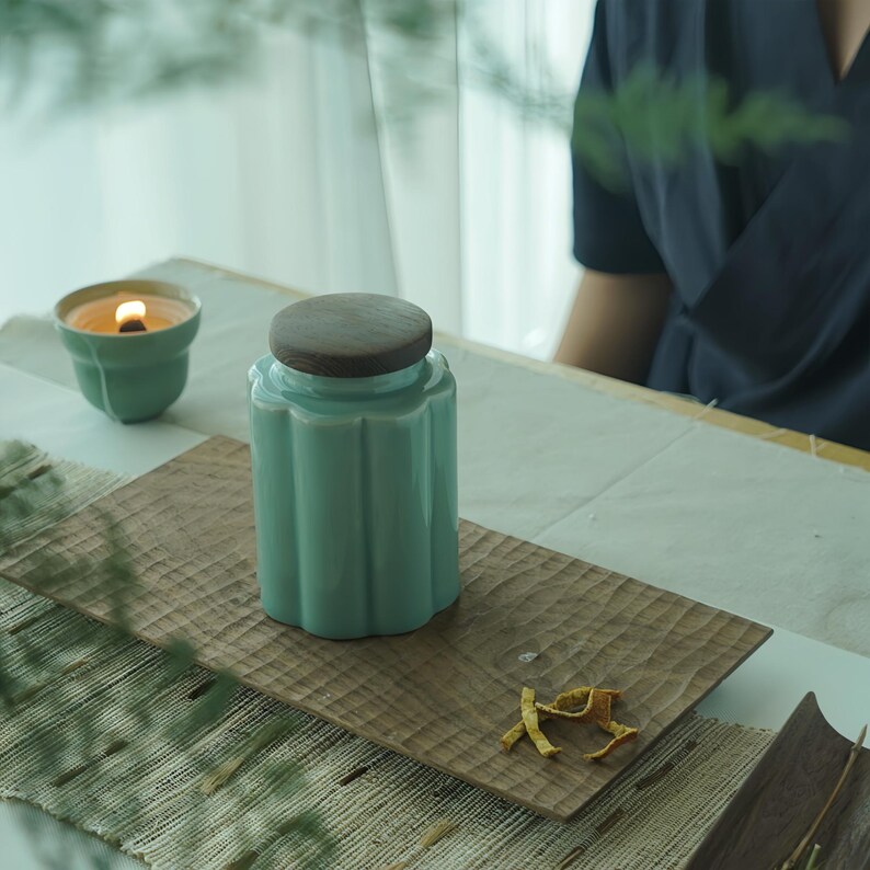 May include: A light teal ceramic jar with a wooden lid sits on a carved wooden tray. A lit teal candle is in the background. The jar has a fluted design and a dark brown lid. The scene is set on a table with a neutral background.
