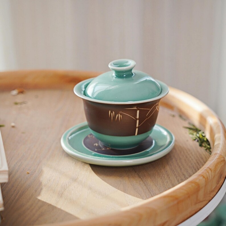 May include: A green and brown ceramic teacup with a lid and a matching saucer. The teacup has a bamboo design on the side.