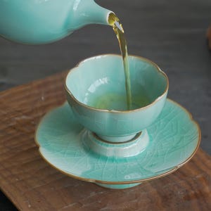 May include: A light mint green ceramic tea cup and saucer set with a delicate crackle glaze finish and gold-toned rims. A matching teapot pours golden tea into the cup, which features a scalloped edge design on both the cup and saucer.